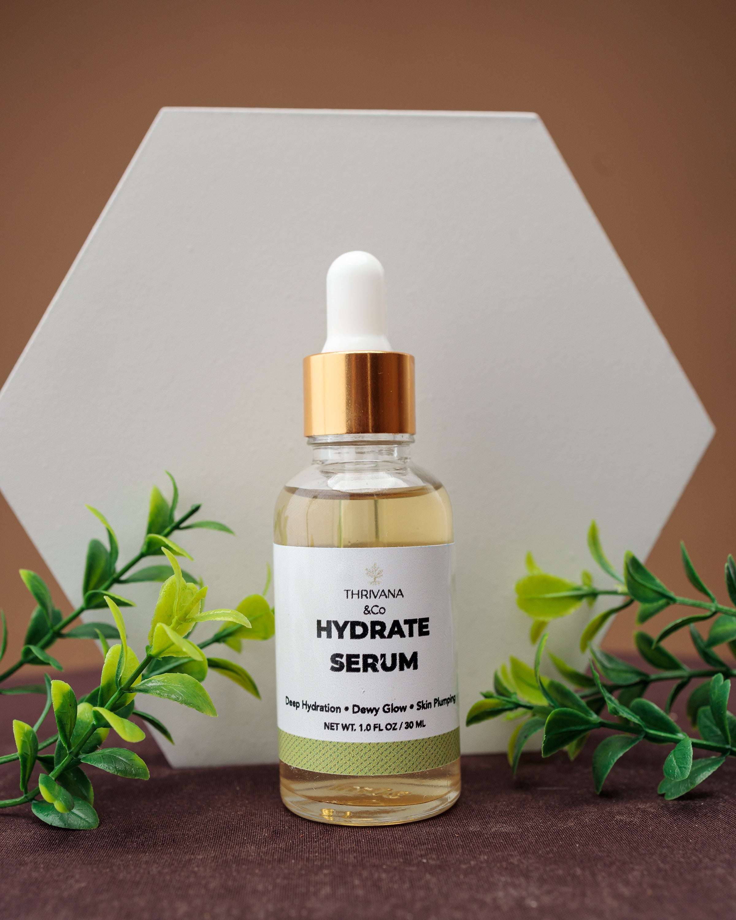 Bottle of Therivana Hydrate Serum with a dropper on a brown background with green leaves.